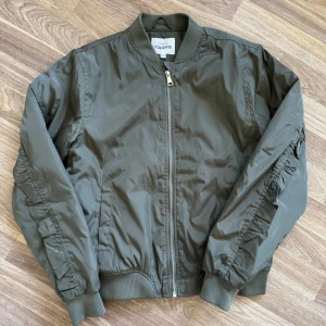 Olivgrön bomberjacka med volang KappAhl - Snygg olivgrön bomberjacka från KappAhl med dragkedja framtill och coola volangdetaljer längs ärmar och rygg. Jackan har ribbade muddar och klassisk bomberkrage. Aldrig använd. Xs