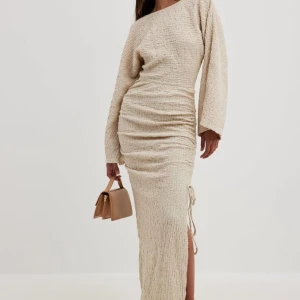 Beige långklänning med slits - Supersnygg beige långklänning med strukturerat tyg, vida långa ärmar och dragsko med slits på sidan. Klänningen har en relaxed fit och markerad midja med rynkade detaljer. Perfekt för dig som gillar en stilren och trendig look. Använd 2 ggr och är från nakd i nyskick nypris 899