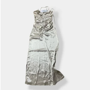 Champagnefärgad satinklänning About You - Lång, champagnefärgad satinklänning med smala axelband och snygg snörning framtill. Klänningen har en glansig finish, figurnära passform och dragkedja i ryggen. Perfekt för dig som vill sticka ut med en elegant och trendig look.