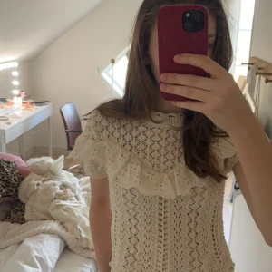 Volangtop från Zara - Supersöt beige stickad blus från Zara med korta ärmar och broderad volangdetalj över bröstet. Blusen har ett genombrutet mönster och ribbad halsringning. Perfekt för vår och sommar när du vill ha en romantisk och trendig look.