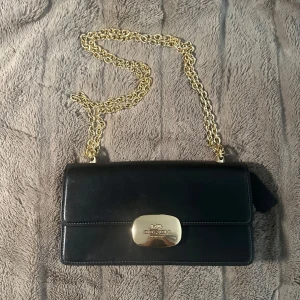 Svart axelväska från Coach i skinn - Stilren svart axelväska från Coach i slätt skinn med guldfärgad kedjerem och elegant logga på metallspännet. Kedjan går att göra kortare eller längre beroende på hur man vill bära väskan. Väskan har ett klassiskt rektangulärt format lock och insida i ljusbrunt material. Perfekt för dig som gillar lyxiga detaljer och tidlös design.