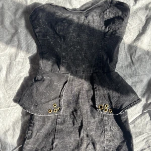 Svart jeansklänning - Svart axelbandslös kortklänning från Terranova med detaljer i midjan och guldfärgade öljetter framtill. Klänningen har figurnära passform, dragkedja bak. Finns även gummi sak vid bysten som håller den uppe. Skriv för fler detaljer!