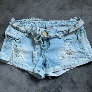Ljusblå lågmidjade jeansshorts - Snygga ljusblå jeansshorts med slitna detaljer och fransad kant. Shortsen har ett blommigt tygbälte och matchande blommigt foder i midjan. 