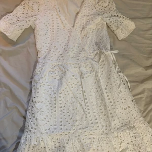 Vit spetsklänning från Vero Moda - Superfin vit kortklänning från Vero Moda med broderade spetsdetaljer och v-ringning. Klänningen har halvlånga ärmar, omlottknytning i midjan och volang nertill. Perfekt för sommarens alla tillfällen!