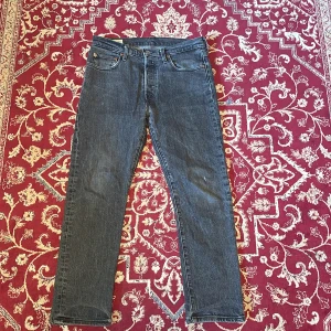 Levi's 501 grå - Klassiska Levi's 501 jeans i mörkblå denim med raka ben och femficksmodell. Jeansen har normal passform och ikonisk läderpatch bak i midjan. Perfekta för dig som gillar tidlös stil och vill ha ett par slitstarka jeans i bomull. Notera en liten slitning på höger knä.