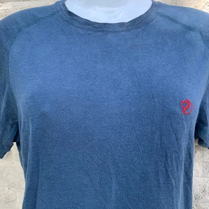 Blå t-shirt från Fjällräven M - Snygg blå t-shirt från Fjällräven med klassisk passform och rund hals. Liten röd Fjällräven-logga broderad på bröstet. Tillverkad i mjuk bomull, perfekt för en chill och avslappnad stil.         Tröjan är gjord av ull. Så både värmer och svalknar beroende på temperatur:)