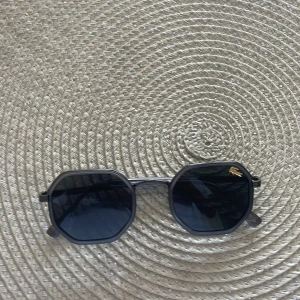 Svarta solglasögon från Lacoste perfekta till somaren! - Snygga svarta solglasögon från Lacoste med åttakantiga bågar och mörka glas. Diskret Lacoste-logga i guld på ena linsen och genomskinliga skalmar med märkets namn på sidan. Perfekt accessoar för att lyfta din stil.