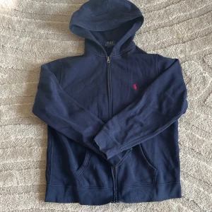 Marinblå hoodie från Polo Ralph Lauren - Marinblå hoodie från Polo Ralph Lauren med dragkedja framtill och klassisk röd logga broderad på bröstet. Tröjan har huva, fickor och långa ärmar. Tillverkad i mjuk bomull, perfekt för chill dagar eller när du vill ha en clean look.