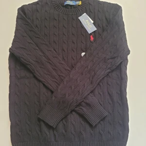 Svart tröja från Polo Ralph Lauren - Kabelstickad svart tröja från Polo Ralph Lauren med rund halsringning och klassisk röd broderad logga på bröstet. Tröjan är långärmad och har ribbade muddar vid ärmslut och nederkant. Perfekt för dig som gillar stilren och tidlös design 😊