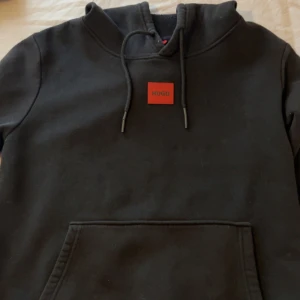 Svart hoodie från Hugo Boss - Svart hoodie från Hugo Boss med röd logotyp framtill. Klassisk modell med huva och snörning, samt stor magficka. Perfekt för dig som gillar stilrena och snygga streetwear-plagg. Mjuk insida och cool design.