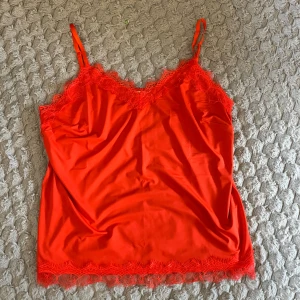 Röd spetslinnetopp från Freequent - Snygg röd linnetopp från Freequent med tunna justerbara axelband och dekorativ spets längs både ringning och nederkant. Toppens material är glansigt och mjukt, vilket ger en lyxig känsla. Perfekt för dig som vill sticka ut med färg och detaljer.