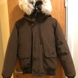 Brun dunjacka med pälskrage Canada Goose - Säljer en brun dunjacka från Canada Goose med stor fluffig pälskant på huvan. Jackan har flera fickor, ribbade muddar och klassisk patch på ärmen. Perfekt för kalla dagar och riktigt snygg streetstil. Finns i Stockholm, Rågsved. Snabb och smidig affär önskas och priset kan diskuteras därefter.