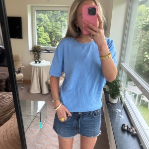 Ljusblå stickad kortärmad tröja - Superfin ljusblå stickad tröja med kort ärm och ribbad nederkant. Tröjan har en enkel, avslappnad passform och en liten metallknapp som detalj framtill. Perfekt till jeansshorts eller kjol för en chill look.