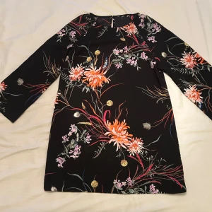 Svart blommig kortklänning - Supersnygg svart kortklänning med färgglada blommor i orange, rosa och lila. Det finns med en extraknapp. Hittar inte storleken men skulle gissa på M