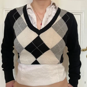 Svart och grå argyle kofta med v-ringning - Snygg croppad kofta med klassiskt argylemönster i svart, grått och vitt. Tröjan har v-ringad hals och långa ärmar. Perfekt för dig som gillar preppy stil och vill sticka ut med en tidlös look. Stickad i mjukt material som känns skönt mot huden.