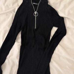 Svart ribbad tröja med dragkedja  - Svart ribbad långärmad tröja från Gina Tricot med half zip och cool ringdetalj på dragkedjan. Slimmad passform och hög krage, perfekt till jeans eller kjol. Superenkel att styla och passar året runt.