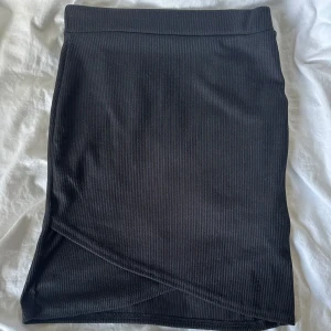 Svart ribbad kjol - Svart tight ribbad kjol med omlott-detalj nertill. Kjolen är kort och har en enkel, stilren design som passar perfekt till crop tops eller oversized tröjor. Materialet är stretchigt och följsamt för en snygg passform