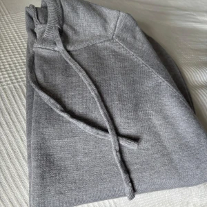 Grå hoodie i merinoull med snörning - Mjuk och stilren grå hoodie med huva och snörning. Tröjan har långa ärmar och ribbade muddar vid ärmslut och nederkant. Helt ny, oanvänd. Storlek XS.