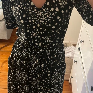 Svart blommig midiklänning med knytband - Svart klänning med vitt blommönster, v-ringning och knytband i midjan. Modellen har korta ärmar. Klänningen är från JACQUELINE de YONG/JDY