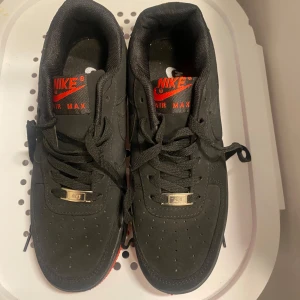 Nike Air Force 1 svart/röd sneakers - Snygga Nike Air Force 1 sneakers i svart mocka med röda detaljer och röd sula. Klassisk låg modell med snörning, perforerad tå och silverfärgad detalj vid snörningen. Perfekta för dig som vill ha en stilren men ändå unik look.