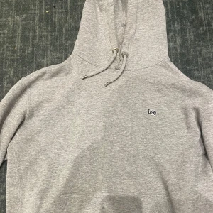 Grå hoodie från Lee med broderad logga - Säljer en stilren grå hoodie från Lee i regular fit. Tröjan har huva med snörning och en liten broderad Lee-logga på bröstet. Perfekt basic-plagg i mjuk bomull som funkar året runt. Passar dig som gillar enkel och clean stil.