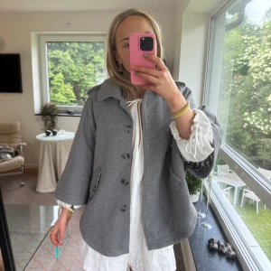 Grå kappa med knappar och fickor - Säljer en grå kappa med bred krage, knappar framtill och två fickor med lock. Jackan har en loose passform och trekvartsärmar. Perfekt för dig som gillar en chill och stilren look. Materialet känns mjukt och är troligtvis bomull eller polyester.
