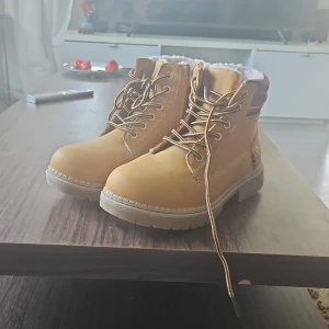 Beige boots med foder och snörning - Snygga beige boots med grov sula, snörning och dragkedja på sidan. Ovandel i skinnliknande material, fodrad med mjuk päls och stickad brun kant upptill. Dekorativt band i rött, vitt och blått på sidan. Perfekta för kyliga dagar.