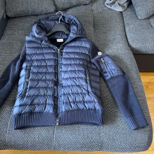 Moncler cardigan - Snygg marinblå pufferjacka från Moncler med stickade ärmar och quiltad front. Jackan har huva, dragkedja framtill och praktisk ficka med dragkedja på ärmen. Perfekt för dig som vill ha en sportig och trendig look under höst och vinter. Pris kan diskuteras vid snabb affär.