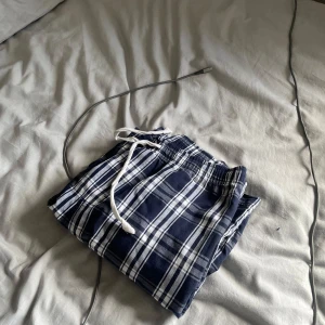 Blårutiga mjukisbyxor med snörning - Säljer ett par blå och vita rutiga mjukisbyxor med elastisk midja och vit snörning. Byxorna är i mjukt bomullstyg och har en avslappnad passform, perfekta för chill dagar hemma.