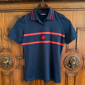 Mörkblå J.Lindeberg golfpike - Snygg mörkblå golfpike från J.Lindeberg med röda ränder över bröstet och på kragen. Klassisk pikékrage, korta ärmar och sportig look. Tillverkad i mjuk och stretchig polyester, perfekt för en aktiv dag på golfbanan.