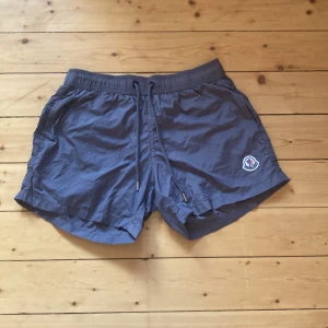 Grå Moncler badshorts med snörning - Snygga grå badshorts från Moncler med elastisk midja och snörning framtill. Shortsen har sidofickor, bakficka med dragkedja och klassisk Moncler-logga på ena benet. Tillverkade i lätt syntetmaterial med meshfoder för extra komfort.
