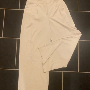 Beiga kostymbyxor från Kappahl - Stilrena beiga kostymbyxor från Kappahl i rak och lite vidare modell. Byxorna har hög midja, pressveck och hällor för bälte. Perfekta för en clean och snygg look. 