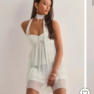 Vit omlott kortklänning med volang - Supersöt vit kortklänning med omlottknytning och volangdetaljer. Klänningen är ärmlös och har en snygg halterneck med scarf-liknande band. Lätt och luftig i ett plisserat tyg. Den är helt oanvänd med prislappen på. Pris kan diskuteras