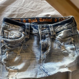 Jeanskjol från Rock Revival - Snygg ljusblå jeanskjol från Rock Revival med slitna detaljer och broderade bakfickor. Kjolen har klassisk femficksdesign, bälteshällor och coola metalldetaljer. Perfekt för dig som gillar en edgy och trendig stil. Helt ny köpt för 1200 