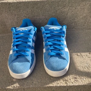 Blå Adidas campus - Snygga blå Adidas campus med klassiska vita ränder. Ovandelen är i mocka och textil, med blå snörning och Adidas logga på hälen. De är i storlek 38 2/3 men passar dom som har 38 i storlek. Perfekta för sommaren och för dig som gillar en stilren look. Skorna kommer dessutom med original boxen!😁