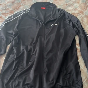 Svart track jacket från Champion XL - Svart track jacket från Champion i storlek XL. Jackan har dragkedja framtill, vita logoband längs ärmarna och broderad Champion-logga på bröstet. Tillverkad i 100% polyester med klassisk sportig look och ståkrage.