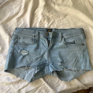 Ljusblå jeansshorts från Lee - Snygga ljusblå jeansshorts från Lee med råa kanter och slitna detaljer framtill. Klassisk femficksmodell med dragkedja och knapp, samt Lee-logga bak på linningen. Perfekta för en chill sommarlook.