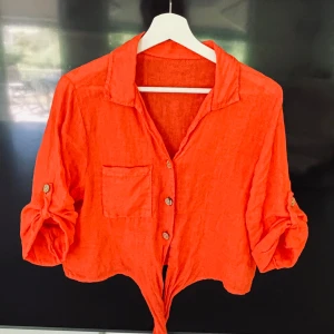 Orangeröd linneskjorta med knyt - Snygg skjorta i 100 % linne med  knyt framtill och 3/4 dels ärm som kan rullas upp och fästas med knapp, klassisk krage och en bröstficka. Perfekt för dig som vill sticka ut med färg och stil.