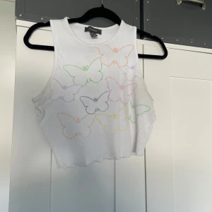 Vit croppad linnetopp med fjärilar - Superfin vit croppad linnetopp från Primark med färgglada fjärilar i pastell på framsidan. Toppen är ribbad, ärmlös och har en vågig kant nertill. Perfekt för sommaren.