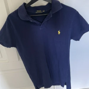 Marinblå pikétröja från Ralph Lauren - Snygg marinblå pikétröja från Polo Ralph Lauren i bomull. Klassisk krage, korta ärmar och gul broderad logga på bröstet.  Custom fit-modell som sitter riktigt bra.