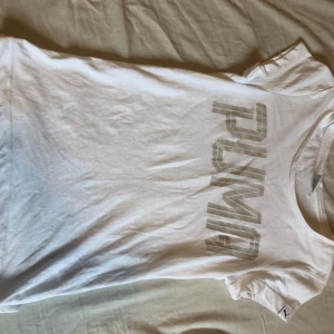 Vit t-shirt från Puma med logga - Vit t-shirt från Puma med stor glansig logga framtill. Klassisk rund halsringning och korta ärmar. Tillverkad i mjuk bomull och har en liten Puma-tag på ärmen. Perfekt för en sportig och clean look.