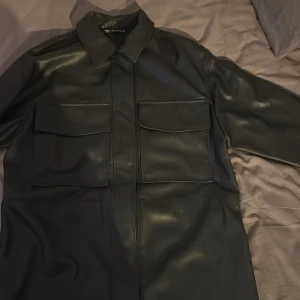 Svart overshirt i skinn från Zara - Snygg svart jacka från Zara i skinnimitation med bröstfickor och klassisk krage. Jackan har raka linjer och stängs med dolda knappar framtill. 