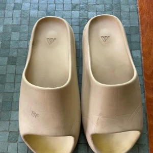 Beige Adidas Yeezy Slides - Beigea Adidas Yeezy Slide sandaler. Perfekta tofflor som är sköna och samtidigt stillsamma. Använde dem förra sommaren vilket man kan se att dem är lite smutsiga men väldigt lätta att tvätta. Extremt bekväma och avslappnade.