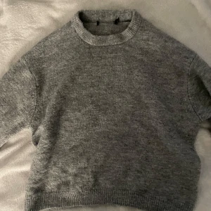 Grå stickad tröja med rund hals - Mysig grå stickad tröja med rund halsringning och ribbade muddar vid ärmslut och nederkant. Perfekt för lager-på-lager och chill dagar. Passformen är normal och tröjan har långa ärmar. Enkel och stilren design som funkar till det mesta.