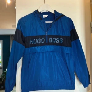 Blå vindjacka från Hugo Boss XS - Snygg blå vindjacka från Hugo Boss med svart panel och stor logga över bröstet. Jackan har huva, lång ärm och half zip framtill. Perfekt för dig som gillar sportig och clean stil. Tillverkad i lätt polyester som känns skön att bära.