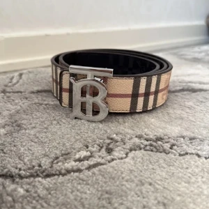 Burberry Bälte - Snyggt bälte från Burberry med klassiskt beige, svart och röd rutigt mönster. Bältet har ett stort silverfärgat spänne med initialerna TB. Perfekt accessoar för att lyfta din outfit.