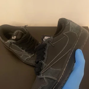 Travis Scott J1 low RÈ🅿️Ş - Svarta Nike Air Jordan 1 Low sneakers i mocka med vita kontrastsömmar och broderade detaljer på hälen. Klassisk rund tå, svart platt sula och snörning. Snygg och stilren modell med ikoniska Air Jordan-loggan och bi-detalj bak.