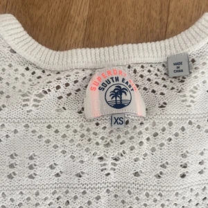 Vit stickad topp från Superdry XS - Snygg vit stickad topp från Superdry i storlek XS. Tröjan har korta ärmar med offshoulder-detalj och ett genomgående hålmönster som ger en luftig och fräsch känsla. Perfekt för varmare dagar och enkel att styla med jeans eller kjol.