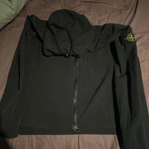 Svart vindjacka från Stone Island med luva  - Svart vindjacka från Stone Island med huva och ikonisk patch på vänster ärm. Jackan har två bröstfickor med knappar och hel dragkedja framtill. Tillverkad i lätt polyester, perfekt för blåsiga dagar. Snygg och stilren design som passar streetwear.