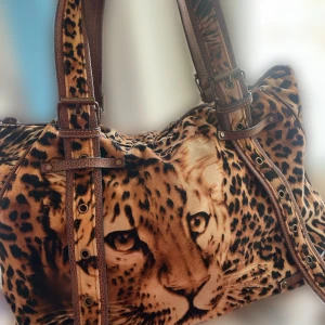 Leopardmönstrad axelväska med detaljer - Stor axelväska med leopardmönster och ett stort tryck av ett leopardansikte. Väskan har bruna remmar med spännen och nitar, samt dragkedja upptill och på baksidan. Materialet är mjukt och känns som sammet. En pärl- och blomnyckelring hänger på ena remmen.